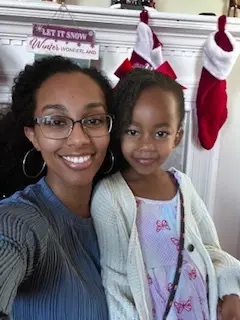 A Black woman smiling into the camera and posing with a four year old Black girl